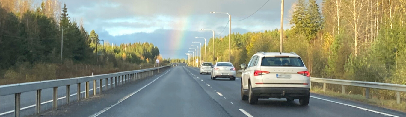 Valkoiset henkilöautot syksyisellä tiellä menevät kohti sateenkaarenpäätä.