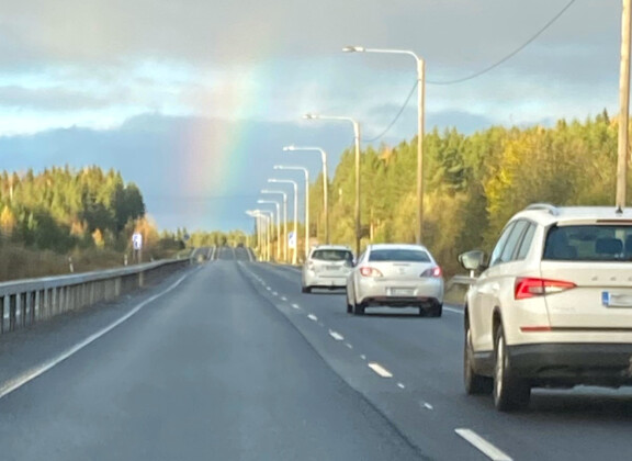 Valkoiset henkilöautot syksyisellä tiellä menevät kohti sateenkaarenpäätä.